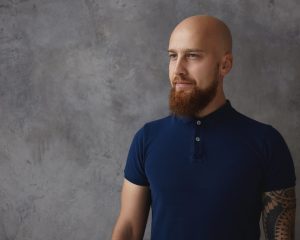 People, style and fashion concept. Studio shot of stylish handsome young Caucasian male with bald head and fuzzy beard standing isolated at blank gray copyspace wall, dressed in trendy polo shirt