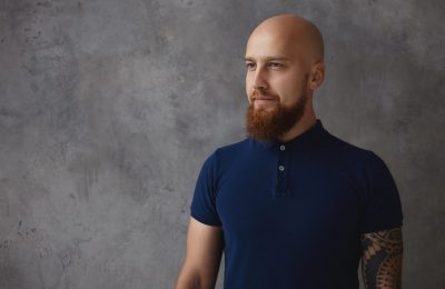 People, style and fashion concept. Studio shot of stylish handsome young Caucasian male with bald head and fuzzy beard standing isolated at blank gray copyspace wall, dressed in trendy polo shirt